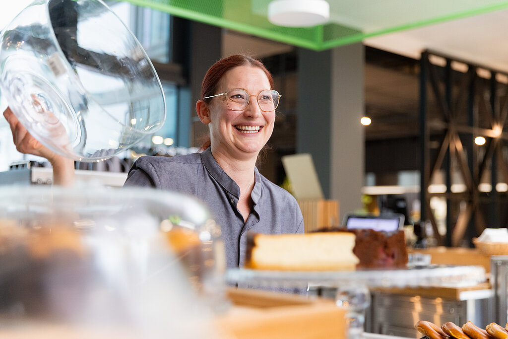 Eine junge Dame/Barista, die die Glashaube einer Etagere hochhebt und einem Kunden einen Käsekuchen zeigt. Sie lacht freundlich.